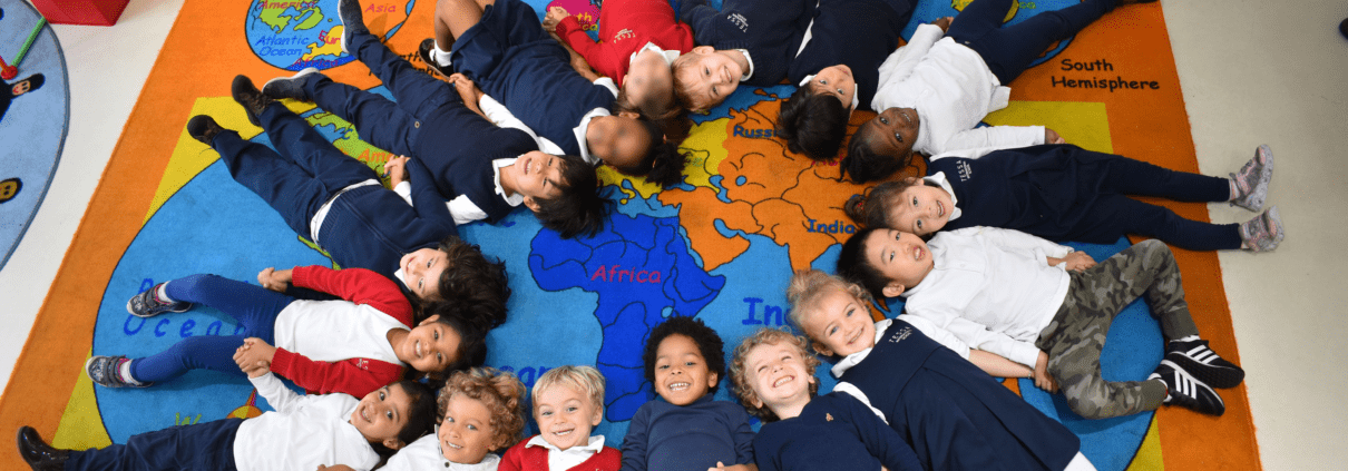 A class of students learning an international school curriculum posing together in a circle on a map carpet in a classroom.