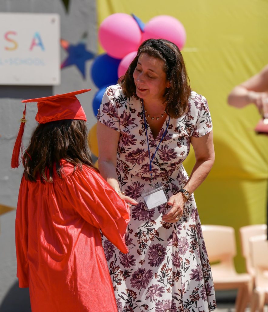 The founder of a school shakes the hand of a student at its kindergarten graduation. The student is wearing a red graduation cap and gown.